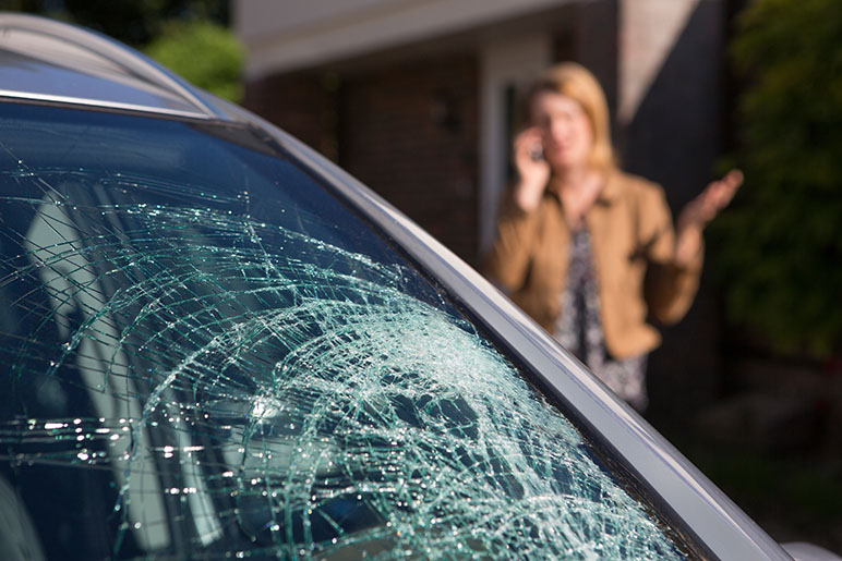 A car that has a cracked windshield and in the distance a woman is on the phone looking stressed