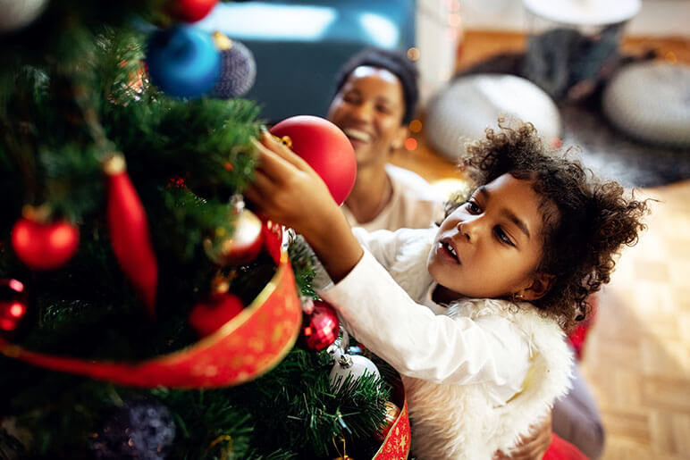 Young girl putting on a Christmas ornament on her Christmas tree while her mom is behind her smiling