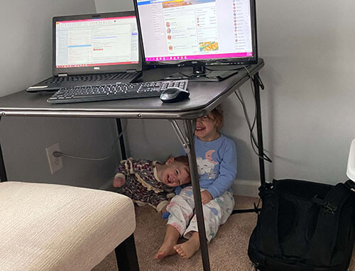 Two smiling children underneath table hiding from their mom who is working from home