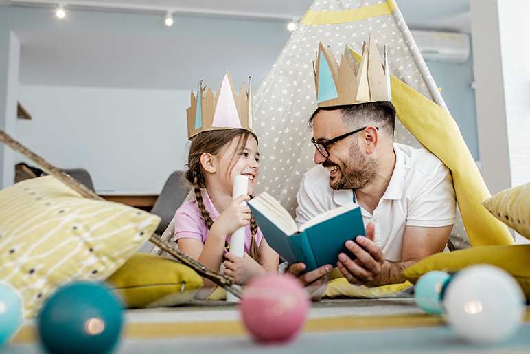 Father and daughter playing on the floor reading a book and wearing paper crowns