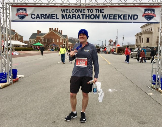 James standing in front of a Carmel Marathon Weekend sign holding his medal up