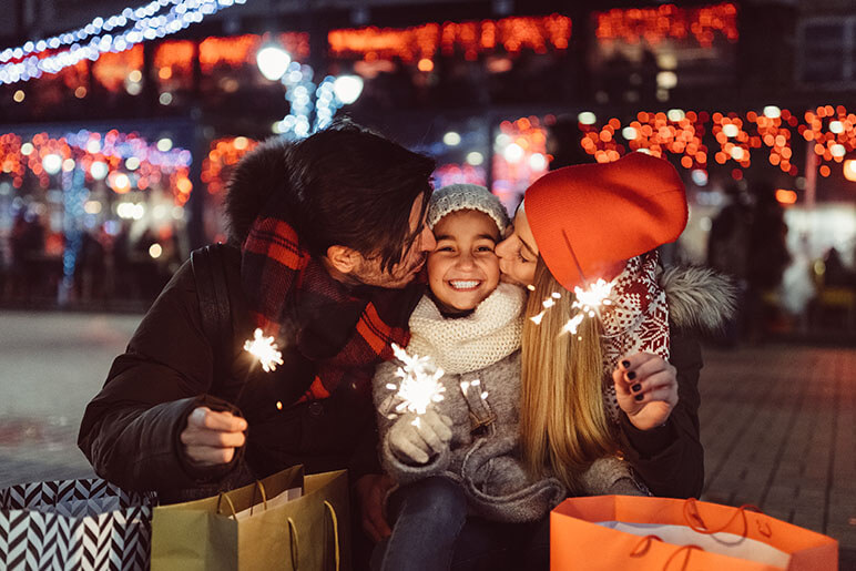 Two parents on both sides of their daughter giving her a kiss on the cheek while holding new year's sparklers