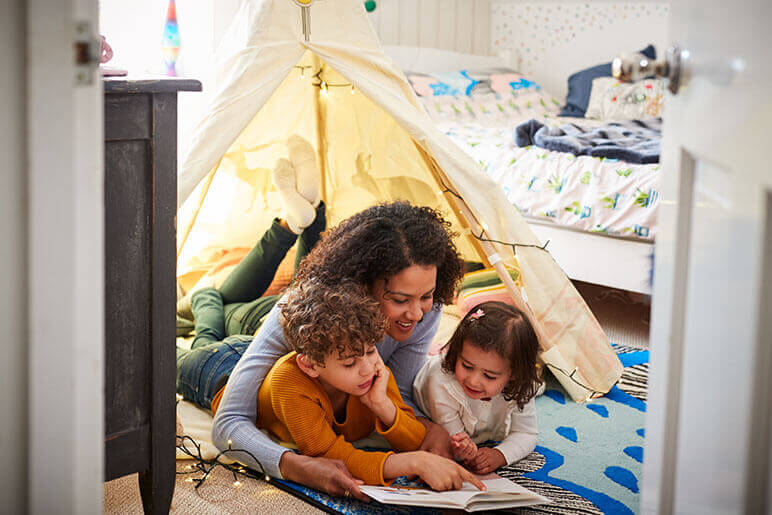 Mom sitting on the ground with young boy and girl reading a book while under a fort