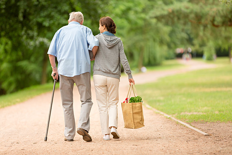 Woman helping an elderly man with a cane walk and carry his groceries through a park