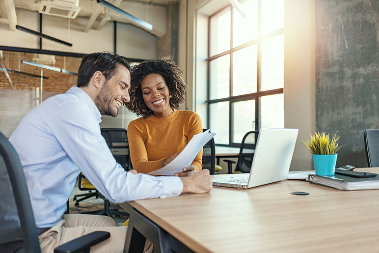 Two young individuals at a desk discussing business looking at paper and a laptop