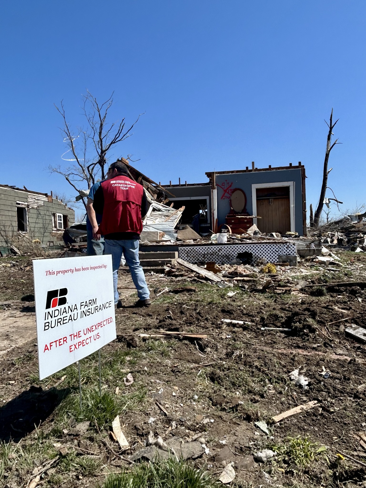 An Indiana Farm Bureau Insurance claims representative stands in the yard of a home destroyed by a tornado in Sullivan, Indiana. In the foreground there is an Indiana Farm Bureau Insurance yard sign that says 